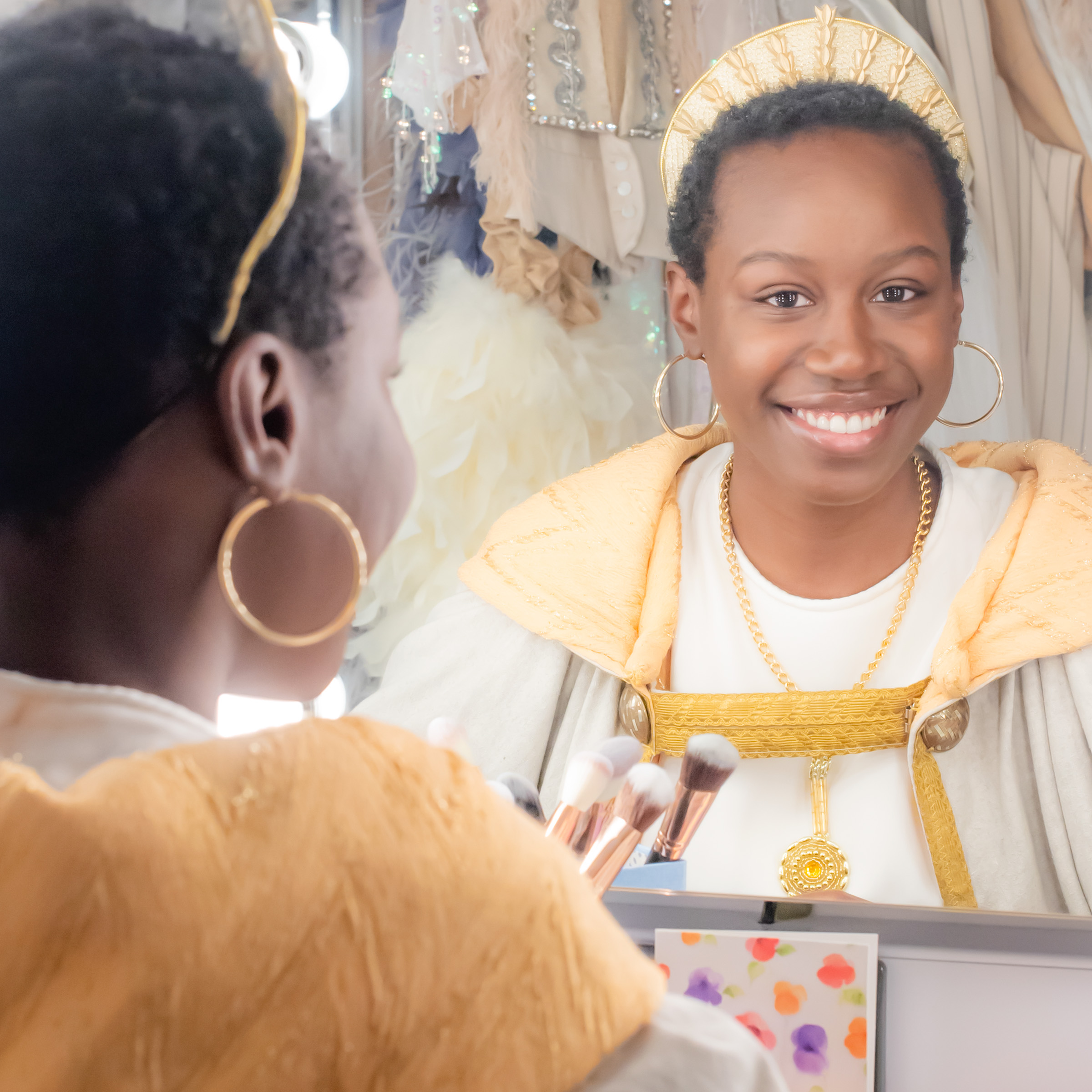 A black women wearing a costume, with a gold headpiece, smiling at herself in the mirror in the dressing rooms.